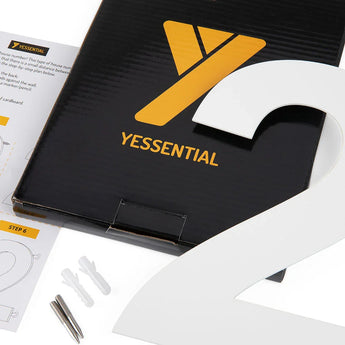 Een zwarte doos met een geel Y logo en "yessential.nl" tekst, een groot Adamo White 30cm huisnummer 2, schroeven, muurankers en instructiebladen zijn gerangschikt op een wit oppervlak.