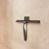 Een Zamoro Gunmetal douchetrekker van yessential.nl met wandhouder en 3M ophangsysteem is bevestigd aan een lichtbeige betegelde muur.