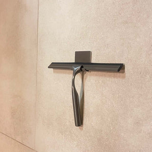 Een Zamoro Gunmetal douchetrekker van yessential.nl met wandhouder en 3M ophangsysteem is bevestigd aan een lichtbeige betegelde muur.