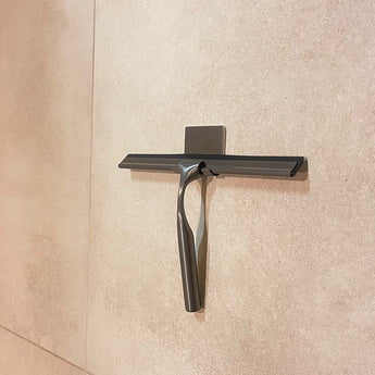 Een Zamoro Gunmetal douchetrekker van yessential.nl met wandhouder en 3M ophangsysteem is bevestigd aan een lichtbeige betegelde muur.
