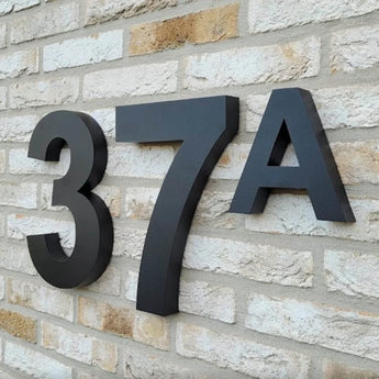 Angelo Black 30cm huisnummer en letter 37A van yessential.nl in moderne stijl, gemonteerd op een lichtgekleurde bakstenen muur.