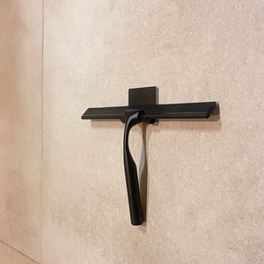 Een Zamoro Black douche trekker van yessential.nl hangt aan een wandhouder met een 3M ophangsysteem tegen een beige betegelde muur.