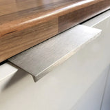 Een close-up van een witte kastdeur met de Enzo RVS handgreep van yessential.nl, met een strakke geborstelde afwerking en een plat rechthoekig ontwerp, afgewerkt met een houten aanrechtblad voor een strakke, moderne look.