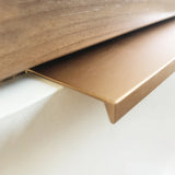 Close-up van Enzo Rose Gold sierstrip van yessential.nl, met een L-vormig profiel en gladde geborstelde afwerking, geïnstalleerd op de rand van een witte muur onder hout - ideaal om te combineren met moderne meubelgrepen voor een stijlvol interieur.