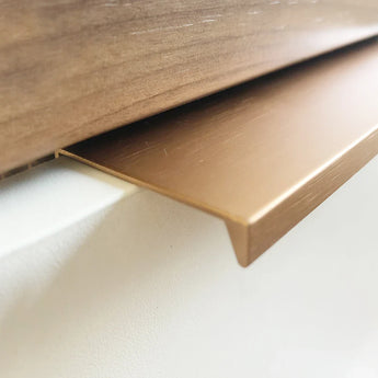 Close-up van Enzo Rose Gold sierstrip van yessential.nl, met een L-vormig profiel en gladde geborstelde afwerking, geïnstalleerd op de rand van een witte muur onder hout - ideaal om te combineren met moderne meubelgrepen voor een stijlvol interieur.