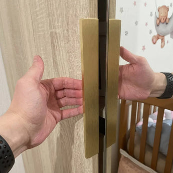 De handen van een persoon reiken om een kledingkast te openen die is uitgerust met Enzo Gold handgrepen van yessential.nl; op de achtergrond verschijnen een houten wieg en een muur met ster- en teddybeerdecoraties.
