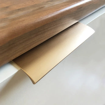 Close-up van een yessential.nl Enzo Gold Deluxe kasttrekhendel bevestigd aan een witte lade, net onder een houten aanrechtblad.