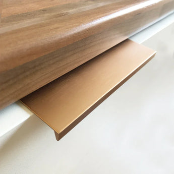 De Enzo Rose Gold handgreep van yessential.nl is een slanke, platte metalen greep die onder een houten aanrecht op een witte kast of lade wordt geïnstalleerd en een moderne touch toevoegt met zijn roségouden afwerking.