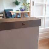 Een close-up van een Enzo RVS schroef van yessential.nl bevestigd in een wit oppervlak, met een onscherpe achtergrond met een tafel, stoelen en kamerplanten in een lichte kamer.