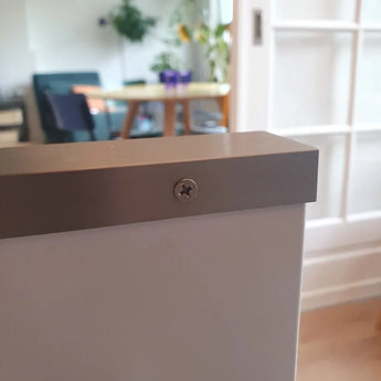 Een close-up van een Enzo RVS schroef van yessential.nl bevestigd in een wit oppervlak, met een onscherpe achtergrond met een tafel, stoelen en kamerplanten in een lichte kamer.