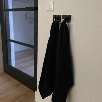 Twee zwarte handdoeken hangen naast elkaar aan Valo Black Hooks van yessential.nl in een minimalistisch interieur, met de haken gemonteerd op een witte muur bij een moderne glazen deur.