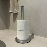 Een yessential.nl Jurado RVS vrijstaande wc-rolhouder staat op een betegelde badkamervloer met twee witte wc-papierrollen. Boven en links van deze stijlvolle toiletaccessoire hangt een beige handdoek.