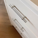 Close-up van twee witte lades met yessential.nl's Arturo RVS handgrepen, getoond onder een hoek. Het strakke Arturo design versterkt een landelijk interieur, tegen een lichte houten vloer.