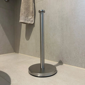 De yessential.nl Jurado RVS vrijstaande wc-rolhouder heeft een roestvrijstalen constructie met een ronde voet en verticale stang, geplaatst op een betegelde badkamervloer. Op de achtergrond hangt een beige handdoek aan de muur.