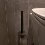 De yessential.nl Roberto Gunmetal verticale toiletrolhouder is gemonteerd op een grijs betegelde muur naast een wit toilet. De lege houder werpt een schaduw op de muur en de vloer.