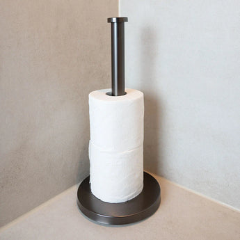 De Delo Gunmetal WC Set (no-drill) van yessential.nl heeft twee toiletpapierrollen en staat in een kamerhoek met lichtgekleurde betegelde muren en oppervlakken.