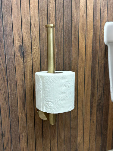 Een rol toiletpapier zit op een verticale messing houder uit de Corrado Gold WC-set van yessential.nl, gemonteerd op een houten wand naast een witte wastafel.