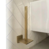 De Bellino Gold toiletrolhouder van yessential.nl, met een modern design en gouden afwerking, heeft een verticale stang en vierkante voet. Hij wordt aan de muur bevestigd op witte diagonale tegels in de buurt van een wit toilet en voegt een minimale stijl toe aan je ruimte.