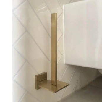 De Bellino Gold toiletrolhouder van yessential.nl, met een modern design en gouden afwerking, heeft een verticale stang en vierkante voet. Hij wordt aan de muur bevestigd op witte diagonale tegels in de buurt van een wit toilet en voegt een minimale stijl toe aan je ruimte.