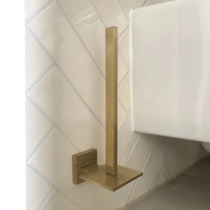 De Bellino Gold toiletrolhouder van yessential.nl, met een modern design en gouden afwerking, heeft een verticale stang en vierkante voet. Hij wordt aan de muur bevestigd op witte diagonale tegels in de buurt van een wit toilet en voegt een minimale stijl toe aan je ruimte.