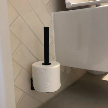 Een rol toiletpapier hangt verticaal aan de Bellino Black reserverolhouder van yessential.nl, bevestigd aan een betegelde badkamermuur naast een wit toilet.