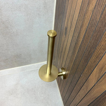 De Roberto Gold toiletrolhouder van yessential.nl, ideaal voor badkameraccessoires, wordt gemonteerd op een houten paneel naast betegelde muren en vloer - hier getoond zonder toiletrol.