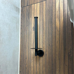 De Roberto Black reserverolhouder van yessential.nl is verticaal gemonteerd op een wand met houtlook panelen, naast lichtgrijze tegels.