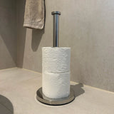 De yessential.nl Ludo Chroom WC Set (no-drill) bevat een roestvrijstalen toiletpapierhouder met twee witte rollen in reliëf op een lichte badkamervloer; een beige handdoek hangt vlakbij. Montage is makkelijk door het zonder boren ontwerp.