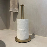 De Ludo Gold WC Set (no-drill) van yessential.nl staat op een licht betegelde badkamervloer met twee toiletrollen en een beige handdoek aan de muur op de achtergrond - allemaal te installeren zonder te boren.