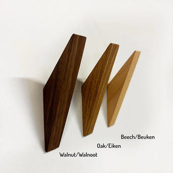 Drie Fabio Beech wandhaken van yessential.nl, in walnoot, eiken en beuken (beukenhout kapstokhaakjes), zijn gemonteerd op een witte muur. Elke minimalistische kapstok heeft een label met de houtsoort in het Engels en Nederlands.