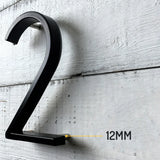 De Amato Black 12,5cm van yessential.nl is gemonteerd op een witte houten muur met een tussenruimte van 12MM, waardoor duidelijk wordt hoe dit moderne zwarte huisnummer afsteekt tegen het oppervlak.
