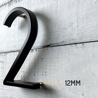 De Amato Black 12,5cm van yessential.nl is gemonteerd op een witte houten muur met een tussenruimte van 12MM, waardoor duidelijk wordt hoe dit moderne zwarte huisnummer afsteekt tegen het oppervlak.
