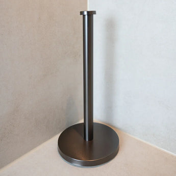 De Jurado Gunmetal papieren handdoekhouder van yessential.nl, met een ronde basis en rechtopstaande stang, staat op een licht aanrechtblad en vult andere Jurado-stukken aan voor een samenhangende look tegen lichtbeige muren.