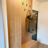 Een moderne houten kapstok met 10x Huso Black haken van yessential.nl houdt een groene jas vast. Ingebouwde kasten en een bankje staan eronder, terwijl een glazen deur op de achtergrond de binnenruimte weerspiegelt.