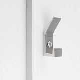 De Huso Chrome kapstokhaak van yessential.nl heeft een geborstelde metallic afwerking, een minimalistisch ontwerp en scherpe hoekige lijnen voor een strakke uitstraling, gemonteerd op een wit oppervlak naast een verticale strip.
