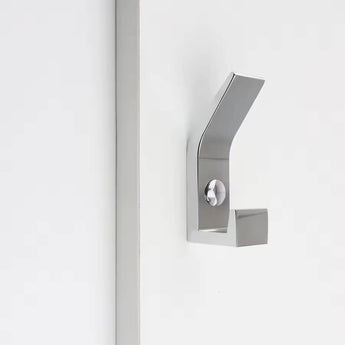 De Huso Chrome kapstokhaak van yessential.nl heeft een geborstelde metallic afwerking, een minimalistisch ontwerp en scherpe hoekige lijnen voor een strakke uitstraling, gemonteerd op een wit oppervlak naast een verticale strip.