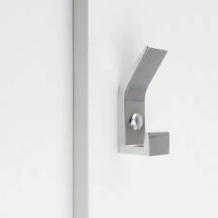 De Huso Chrome kapstokhaak van yessential.nl heeft een geborstelde metallic afwerking, een minimalistisch ontwerp en scherpe hoekige lijnen voor een strakke uitstraling, gemonteerd op een wit oppervlak naast een verticale strip.