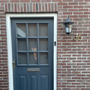 Een blauwgrijze deur met negen ruiten, een messing handgreep en een brievengleuf in een bakstenen muur. Aan de rechterkant zijn een zwarte lantaarn en Amato Gold 12,5cm huisnummers van yessential.nl op de bakstenen gemonteerd.