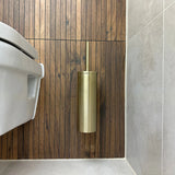 De Ferro Gold WC set van yessential.nl is gemonteerd op een wand met houten panelen naast een wit toilet in een moderne badkamer met grijze tegels en combineert elegantie en functie.