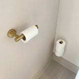 Een bijna lege toiletrol hangt aan de Lorenzo Gold WC set van yessential.nl, bevestigd aan een beige muur. Twee reserverollen liggen op de vloer, als aanvulling op de sobere toiletaccessoires in deze ruimte.