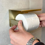 Een persoon met een zilveren horloge plaatst toiletpapier op de Cano Gold WC Set (no-drill) van yessential.nl, gemonteerd op een betegelde muur.