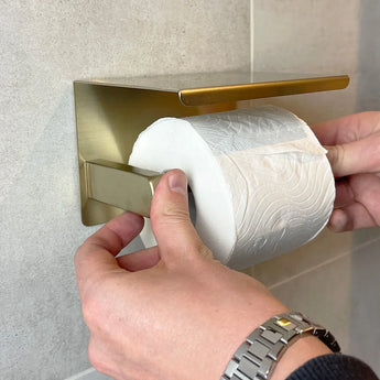 Een persoon met een zilveren horloge plaatst toiletpapier op de Cano Gold WC Set (no-drill) van yessential.nl, gemonteerd op een betegelde muur.