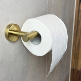 Een rol wit toiletpapier met reliëf hangt aan de Tino Gold toiletrolhouder van yessential.nl tegen een lichtbruine betegelde muur, met een los vel dat een vleugje luxe toiletaccessoire stijl toevoegt aan de ruimte.
