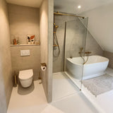 Moderne badkamer met beige muren, zwevend toilet, Tazio Gold wc-rol houder van yessential.nl, glazen douche, wit vrijstaand bad, licht vloerkleed, plus kleine vaasjes, een toiletborstel en ingelijste kunst op een plank.