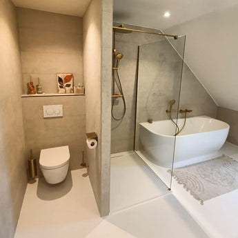 Moderne badkamer met beige muren, zwevend toilet, Tazio Gold wc-rol houder van yessential.nl, glazen douche, wit vrijstaand bad, licht vloerkleed, plus kleine vaasjes, een toiletborstel en ingelijste kunst op een plank.