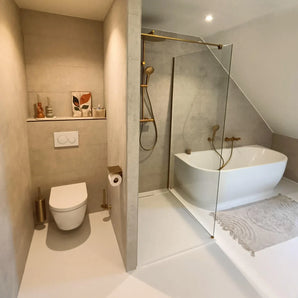 Modernes Badezimmer mit beigen Wänden, schwimmender Toilette, Tazio Gold Toilettenpapierhalter von yessential.de, Glasdusche, weißer freistehender Badewanne, hellem Teppich sowie kleinen Vasen, einer Toilettenbürste und gerahmter Kunst auf einem Regal.
