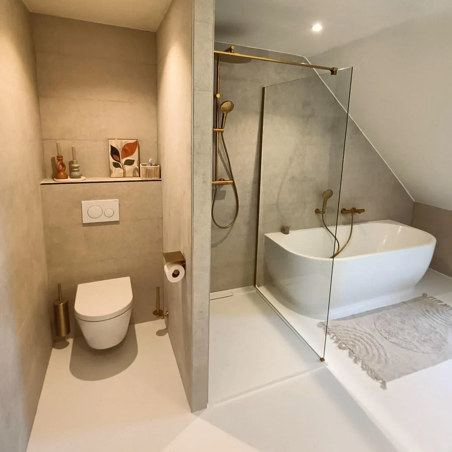 Moderne badkamer met beige muren, zwevend toilet, Tazio Gold wc-rol houder van yessential.nl, glazen douche, wit vrijstaand bad, licht vloerkleed, plus kleine vaasjes, een toiletborstel en ingelijste kunst op een plank.