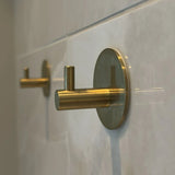 Een close-up van twee Rudro Gold handdoekhaken van yessential.nl met ronde basis en cilindrisch ontwerp, zelfklevend gemonteerd op een lichtgekleurde tegelwand-de ideale oplossing zonder boren.