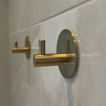 Een close-up van twee Rudro Gold handdoekhaken van yessential.nl met ronde basis en cilindrisch ontwerp, zelfklevend gemonteerd op een lichtgekleurde tegelwand-de ideale oplossing zonder boren.
