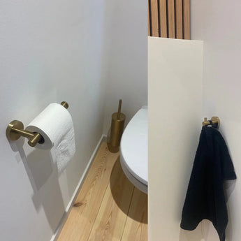 Een kleine moderne badkamer met een licht houten vloer, een wit toilet, gouden toiletpapierhouder en borstel, en de Justo Gold Hook van yessential.nl die een zwarte handdoek vasthoudt voor een elegante touch.